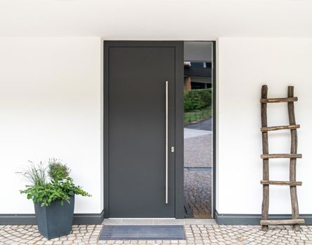 Porta d'ingresso moderna di colore grigio scuro, con design a filo muro, maniglia verticale in acciaio inox e elemento in vetro