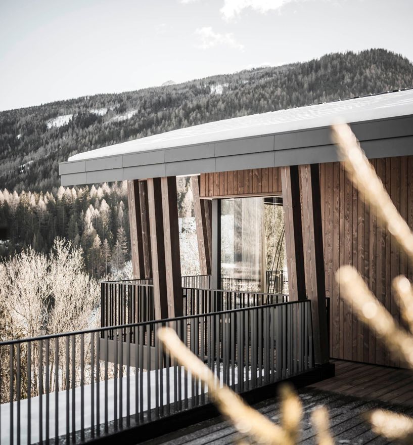 Holzfassade mit modernen Balkonen vor verschneitem Wald und Berglandschaft im Hintergrund.