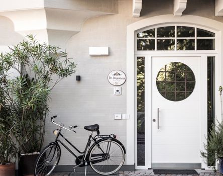 Ingresso bianco con porta rotonda in vetro, piante in vaso e bicicletta su facciata grigia.